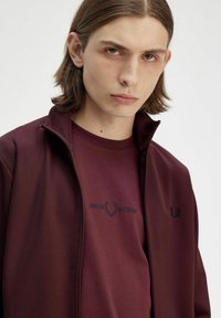 Giacca zip-up borgogna realizzata in tessuto liscio, con logo "Fred Perry" ricamato e t-shirt girocollo abbinata sotto.