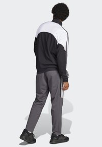 Tuta sportiva con parte superiore nera e bianca con colletto alto, pantaloni grigi con strisce laterali e sneakers nere.