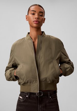 Frau trägt eine olivgrüne Bomberjacke mit den Händen in den Taschen, kombiniert mit schwarzen Jeans vor einem schlichten grauen Hintergrund.