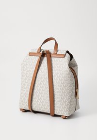Sac à dos en tissu crème avec un motif MK répétitif, avec des bretelles et des accessoires en cuir marron, une poche latérale zippée et une forme structurée.