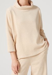 Pull en maille côtelée beige avec un col roulé, des manches trois-quarts et une coupe décontractée, fabriqué dans un matériau doux et texturé.