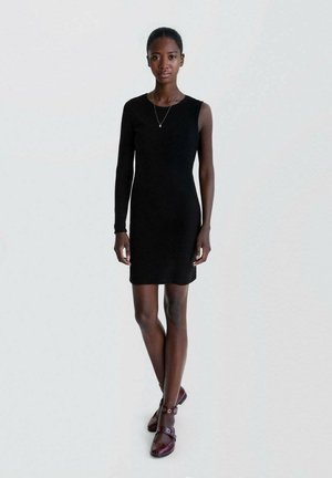 Black ribbed dress with one long sleeve, fitted design. Paired with dark red shoes featuring round accents and ankle straps.
