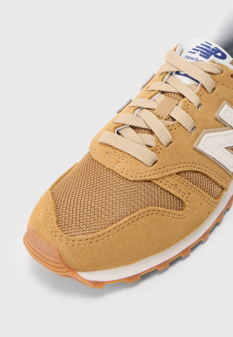 Baskets beiges présentant une combinaison de daim et de matériau en maille, avec des lacets beige et un logo blanc accentué par une semelle en caoutchouc texturée.