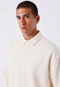Jonge man met kort haar die een crèmekleurig poloshirt draagt met een klein PXG-logo op de borst en een ringoorbel in zijn linkeroor.