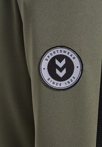 Rond zwart-wit label met de tekst "Sportswear Since 1923" en twee naar beneden wijzende pijlen, genaaid op olijfgroen en zwart stof.