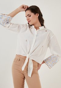 Blouse en lin blanc avec un devant noué, ornée de broderies bleues et dorées sur des manches évasées. La chemise a un col et des boutons sur le devant.
