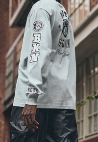 Helles graues Langarmshirt mit grafischem Text und Logos, einschließlich "BKN" und "Brooklyn", kombiniert mit schwarzen Cargo-Hosen.