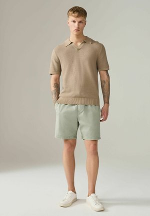 Mladý muž s tetovaniami, oblečený v béžovom pletenom krátkym rukáve polu, svetlozelených šortkách a bielych teniskách, stojaci na jednoduchom pozadí.