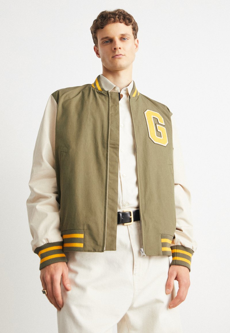 Chaqueta bomber verde oliva con puños y cuello a rayas amarillas, que presenta un gran parche blanco con la letra "G". Lleva puesta sobre una camisa de color claro.