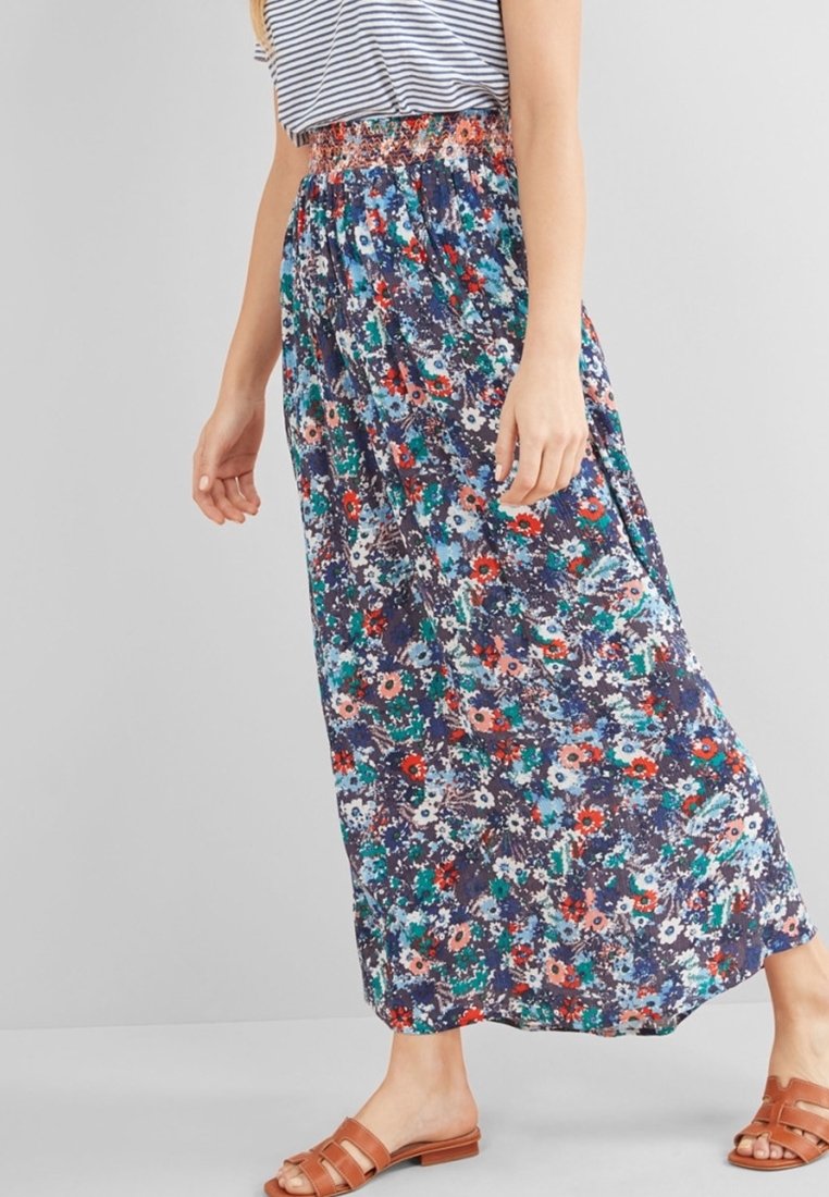 Femme portant une longue jupe maxi blue à motifs floraux, un haut rayé rentré et des sandales brunes à enfiler, se tenant devant un fond uni.