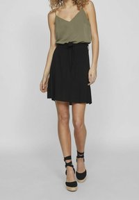 Olivgrön ärmlös topp med v-ringning, kombinerad med en svart högmidjad kjol med ett bindebälte. Svarta espadrilloskor med ankelremmar.