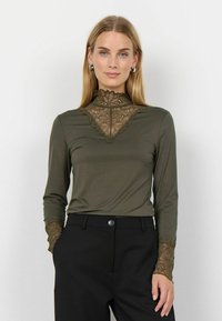 Maglia a maniche lunghe verde oliva con un alto colletto in pizzo e dettagli in pizzo alle maniche, abbinata a pantaloni neri a vita alta.
