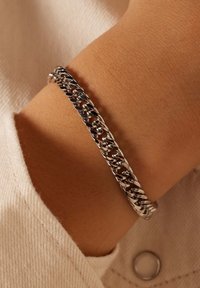 Zilveren schakelarmband, met in elkaar grijpende schakels en een gepolijste afwerking. Draagbaar om een pols, met een gestructureerde lichte stof op de achtergrond.