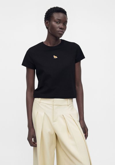 Γυναίκα που φοράει μαύρο κοντομάνικο μπλουζάκι με μικρό κεντημένο τίγρη και κρεμ πλισέ παντελόνι ψηλόμεσο με φαρδιά πόδια.