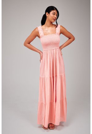 Jonge vrouw met donker haar draagt een mouwloze roze maxi-jurk met gerimpelde bandjes, staand met handen in haar zij tegen een effen achtergrond.