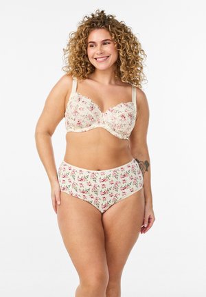 Smilende kvinde med krøllet hår iført cremefarvet blomstret blondebh og matchende højttaljede trusser, stående mod en hvid baggrund.