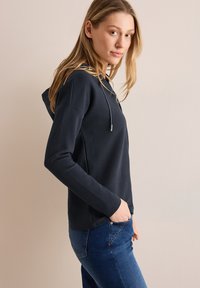 Mørkeblå hættetrøje med ribbet tekstur, snoretræk og sidelommer, parret med blå jeans i en sideprofil.