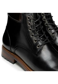 Černé kožené kotníkové boty s kulatou špičkou, šněrováním, kovovými průchody a kontrastní hnědou dřevěnou podrážkou. Hladká textura.