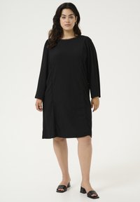 Vestido negro de longitud hasta la rodilla con mangas largas, tejido texturizado, escote redondeado y costuras decorativas a lo largo de los lados. Combinado con sandalias negras.