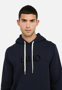 Jeune homme aux cheveux courts et foncés portant un sweat à capuche bleu marine avec des cordons blancs et le mot "Liu Jo" sur la poitrine.