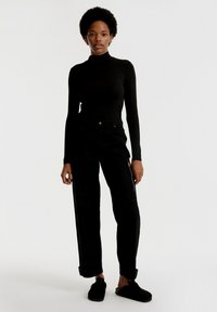 Zwarte lange mouwen turtleneck top gecombineerd met hooggewaisted zwarte broeken met zijritsen. Model draagt zwarte instap schoenen.