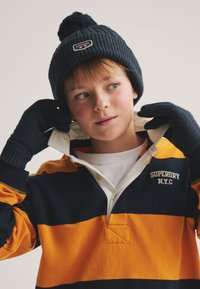 Donkerblauwe gebreide muts met pompon, bijpassende handschoenen en een gestreept oranje en marineblauw polohemd met lange mouwen en een witte kraag.