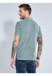 Green polo shirt with short sleeves, textured fabric, and a small logo detail at the back. Worn with blue jeans.