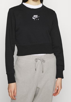 Persoon draagt een zwarte Nike Air cropped sweatshirt over een wit shirt en lichtgrijze joggingbroek met trekkoord.