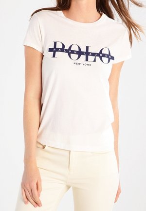 Vrouw draagt een wit T-shirt met korte mouwen met het logo "Polo Ralph Lauren New York" in marineblauw, gecombineerd met lichtbeige broek.