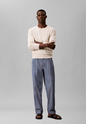 Mann steht mit verschränkten Armen, trägt ein beiges Langarmshirt, blaue lockere Hosen und schwarze Sandalen vor einem schlichten grauen Hintergrund.