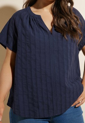 Kobieta ubrana w granatową teksturowaną bluzkę z krótkim rękawem i dekoltem w kształcie litery V oraz w niebieskie dżinsy, stojąca z jedną ręką na biodrze.