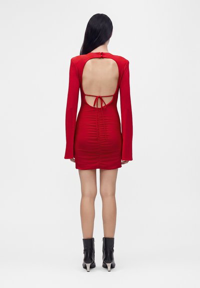 Femme aux longs cheveux noirs portant une mini-robe rouge à manches longues avec un dos ouvert et des bottines noires, debout sur un fond blanc.