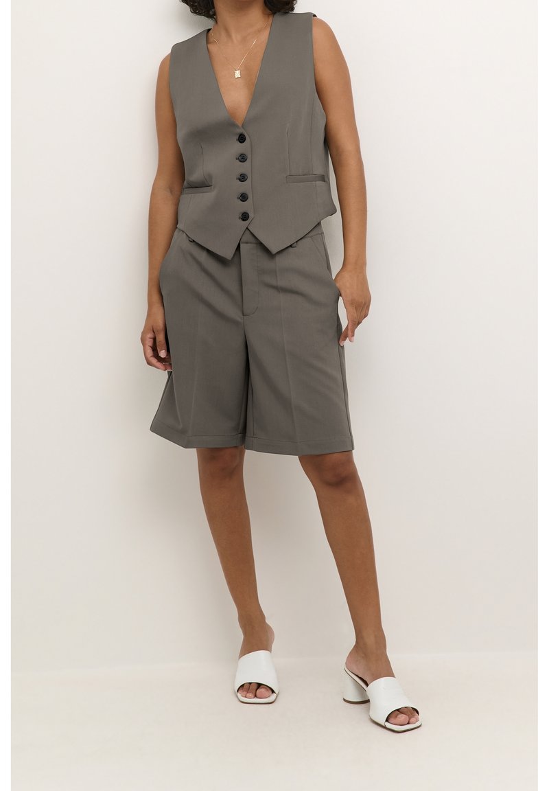 Femme portant un gilet gris boutonné et un short long assorti avec des mules blanches ouvertes à talons, debout devant un fond clair uni.