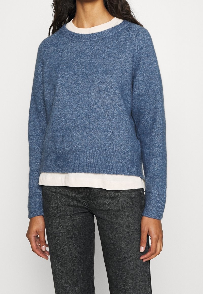 Femme portant un pull en tricot bleu superposé à une chemise blanche, associée à un jean gris foncé, debout devant un fond uni.