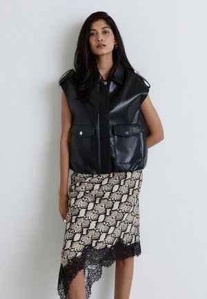 Femme aux longs cheveux foncés portant un gilet en cuir noir sans manches et une jupe asymétrique à motif serpent avec une bordure en dentelle noire.