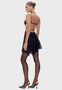 Kobieta w czarnej sukience na ramiączkach z przezroczystą spódnicą, czarnych rajstopach z szwem i czarnych szpilkach, pozująca z boku na jednolitym tle.
