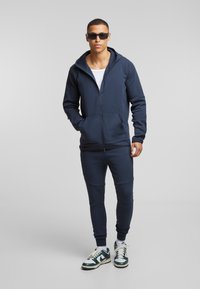 Mornarska zip-up dukserica s prednjim džepom, u kombinaciji s odgovarajućim trenirkama. Teksturirana tkanina, atletski kroj, uparen s zelenim i bijelim tenisicama.