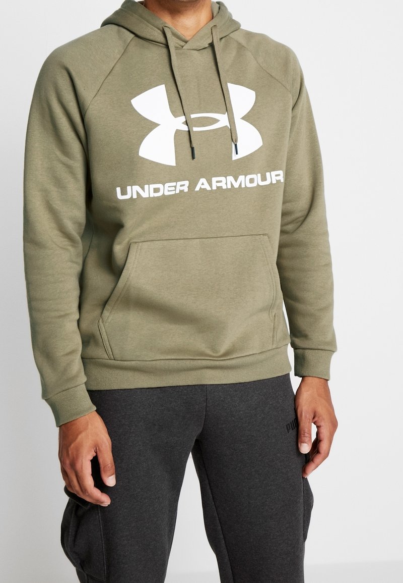 Homme portant un sweat à capuche Under Armour vert olive et un pantalon de survêtement Puma gris foncé, debout devant un fond clair uni.