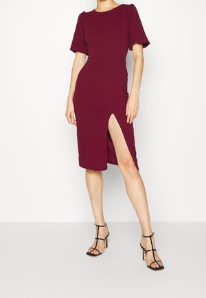 Kvinde iført en tætsiddende, knælænge bourgognefarvet kjole med puffede korte ærmer og et højt slids på låret, kombineret med sorte, remmeklædte hæle.