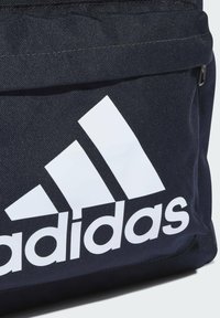 Zaino in tessuto blu navy con un grande logo bianco Adidas e tre strisce diagonali. La texture è resistente con una finitura liscia.