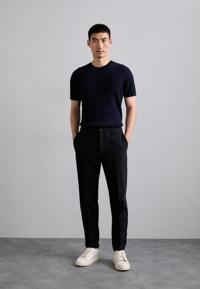 Camisa de manga corta de canalé en color navy, pantalones negros ajustados con pliegues en la parte delantera y zapatillas blancas. El modelo está de pie con las manos en los bolsillos frente a un fondo gris.