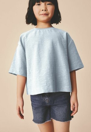 Kind mit kurzem schwarzem Haar trägt hellblaues, strukturiertes, übergroßes Hemd und dunkle Jeansshorts, steht vor neutralem Hintergrund.