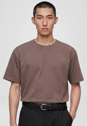 Jeune homme aux cheveux noirs courts portant un t-shirt marron rentré dans un pantalon noir avec une ceinture noire tressée sur un fond clair uni.