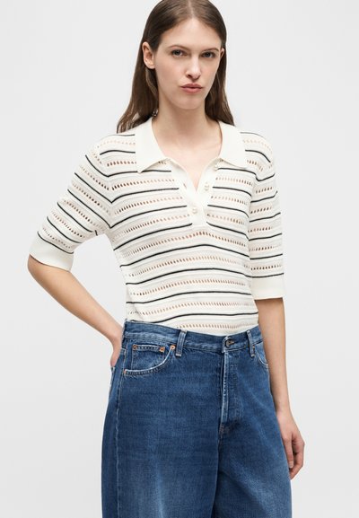 Femme portant un polo en maille blanc à rayures avec des manches trois-quarts et un jean bleu taille haute, debout avec une main sur la hanche.