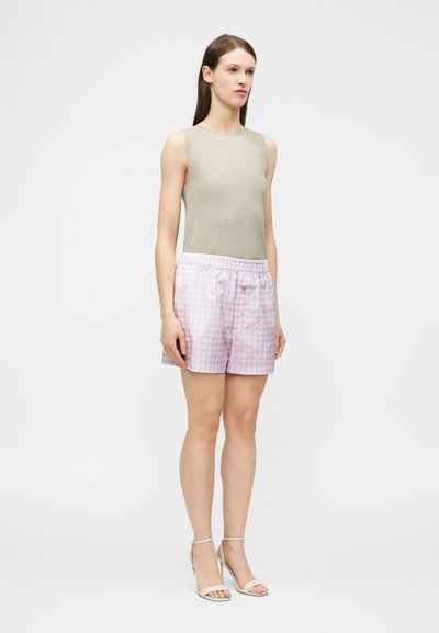 Femme portant un haut beige côtelé sans manches, un short vichy rose et blanc avec des boutons, et des sandales blanches à talons hauts, debout devant un fond blanc.