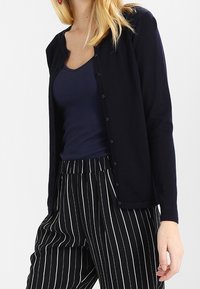 Marinefarbener Cardigan aus weichem Material mit Rundhalsausschnitt, langen Ärmeln und Knopfverschluss, kombiniert mit gestreiften Schwarz-Weiß-Hosen.