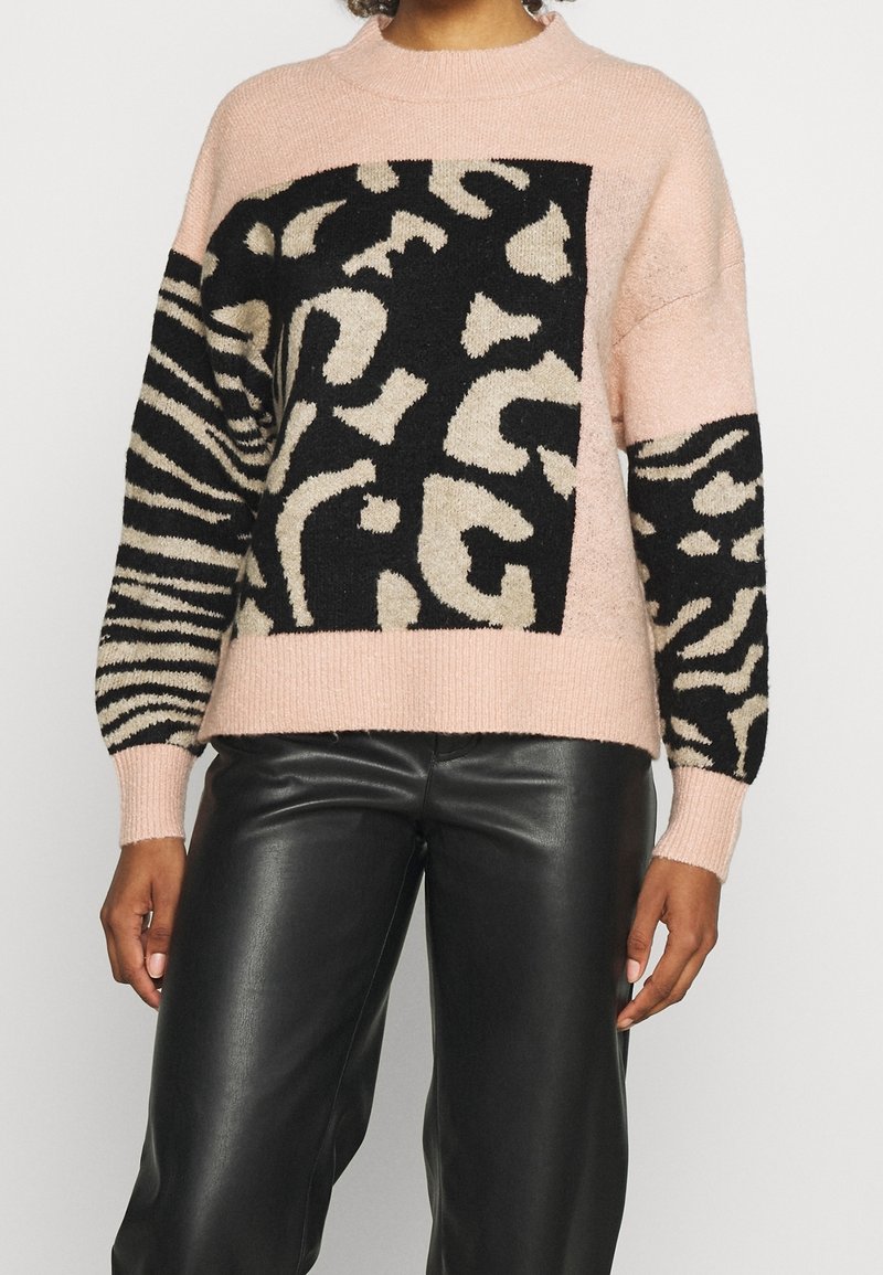 Femme portant un pull rose avec des motifs abstraits d'imprimé animal en noir et beige, associé à un pantalon en cuir noir.