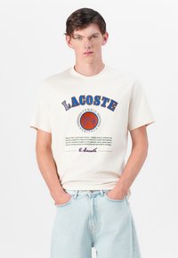 Krēmkrāsas kokvilnas T-krekliņš ar zilu un oranžu 'LACOSTE' logo, tenisa grafiku un drukātu tekstu. Īsas piedurknes, klasiskā piegriezuma.