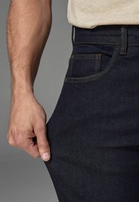 Jeans de mezclilla oscura con un ajuste tapered, que presentan costuras en contraste, un bolsillo frontal y una mano agarrando la tela cerca del muslo.