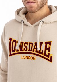 Vajszínű kapucnis pulóver, húzózsinórral, nagy bordó "Lonsdale" logóval és alatta a "London" felirattal. Puha anyagú, laza fazonú.